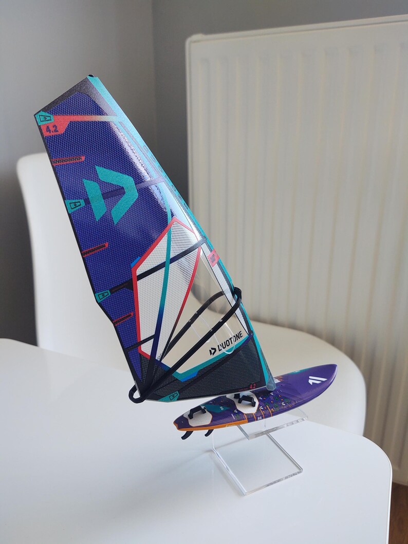 May include: A miniature windsurfing sail and board set. The sail is primarily blue with teal and red accents, and the board is purple. The sail features the text "DUOTONE" and the number "4.2". The set is displayed on a clear stand.