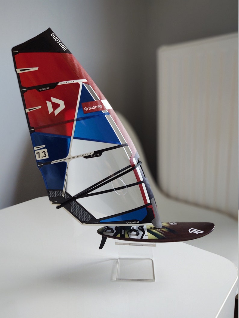 Puede incluir: Una vela de windsurf roja, blanca y azul con el logotipo de Duotone y el texto "E-Type" y "7.3". La vela est&aacute; montada en un soporte de acr&iacute;lico transparente.