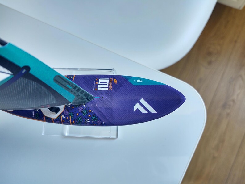 May include: A blue and purple windsurf board with the word "ULTRA" printed on it. The board has a white logo and the number "40" printed on it. The board is on a clear stand.
