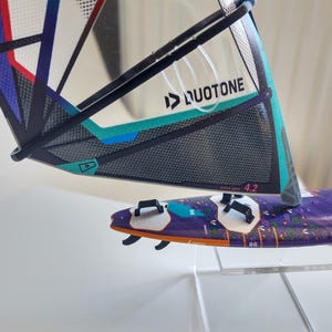 May include: A windsurfing board and sail set. The sail is black, teal, and purple with the Duotone logo. The board is purple with white and black accents. The sail reads "SUPER HERO 4.2".