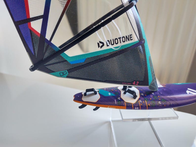 May include: A windsurfing board and sail set. The sail is black, teal, and purple with the Duotone logo. The board is purple with white and black accents. The sail reads "SUPER HERO 4.2".