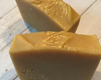 May include: Two bars of handmade soap with a light brown color and a glossy finish. The top bar has a decorative swirl pattern, and the bottom bar has a duck stamp. The soap is rectangular and appears to be freshly cut.