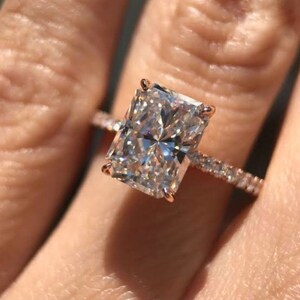 May include: A rose gold engagement ring with a large, rectangular, clear diamond. The diamond is set in a prong setting and the band is a pave setting with smaller diamonds.
