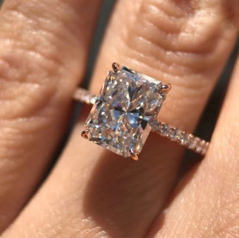 May include: A rose gold engagement ring with a large, rectangular, clear diamond. The diamond is set in a prong setting and the band is a pave setting with smaller diamonds.