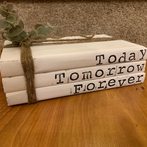 Puede incluir: Una pila de tres libros de madera blancos atados con hilo y adornados con una ramita de vegetación. Los libros están estampados con las palabras "Today", "Tomorrow" y "Forever" en una fuente negra de estilo máquina de escribir. El conjunto está sobre una superficie de madera.