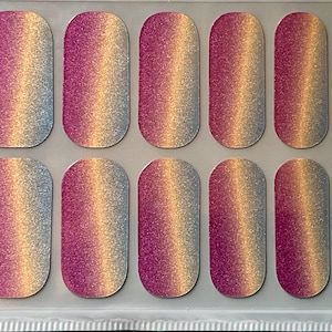 May include: A sheet of nail polish strips with a gradient design. The strips transition from a silver-blue to a gold and then to a magenta color. The nail strips are rectangular with rounded edges.