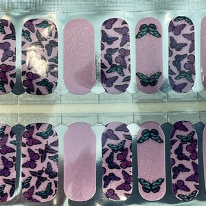 May include: Nail strips featuring a butterfly design in shades of purple and teal on a light pink background. The nail art strips have a mix of butterfly patterns and solid color sections. The strips are packaged on a silver backing.