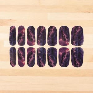Op de afbeelding: Een set nagelwraps met een marmerpatroon. De wraps hebben een donkerpaarse basis met wervelende roze en witte patronen, die doen denken aan een melkwegstelsel. De nagelwraps zijn gerangschikt op een licht houten oppervlak.