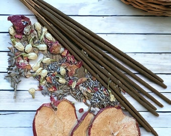 May include: Brown incense sticks, dried apple slices, and a mix of dried flowers and herbs, such as rose petals and lavender, are arranged on a white wooden surface. A wicker basket filled with dried fruit is in the background.