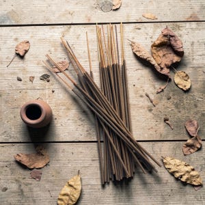 Puede incluir: Una colección de varillas de incienso con puntas doradas, dispuestas sobre una superficie de madera rústica. Una pequeña olla de color terracota y hojas secas están dispersas alrededor del incienso, creando una estética natural y terrosa. Las varillas de incienso son marrones.