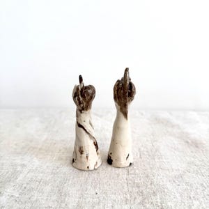 May include: Two ceramic sculptures of hands with the middle finger raised. The sculptures are white with brown accents and sit on a neutral-colored surface. The hands are the focal point, conveying a bold statement.