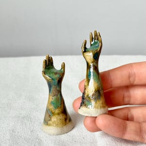 May include: Two ceramic hand sculptures with open palms. The hands are glazed with a mix of green, brown, and gold colors. The base of each hand is a light beige color. The sculptures are displayed on a white surface.