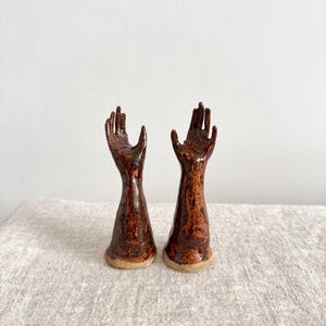 May include: Two ceramic hand sculptures with a brown glaze. The hands are in a gesture of offering or receiving, with fingers slightly curved. The bases are a lighter brown, and the background is a neutral color.