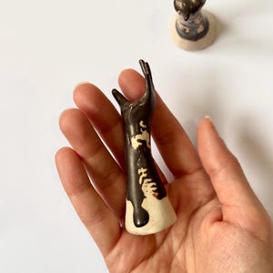 May include: A ceramic sculpture of a hand with a dark brown glaze over a cream base. The hand is raised in a gesture, with the fingers slightly curved. A second, similar sculpture is in the background.