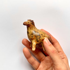 May include: A small, handcrafted ceramic horse figurine. The horse is mainly tan with brown and black markings, and has a textured surface. The figurine is held in a hand against a white background.
