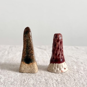 May include: Two ceramic incense holders. One is brown and gold with a textured surface, the other is red with a textured surface and a white base. Both are cone-shaped and sit on a neutral fabric.