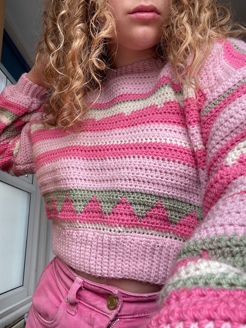 May include: A pink and green striped crocheted sweater with a geometric pattern. The sweater is cropped and has a relaxed fit.