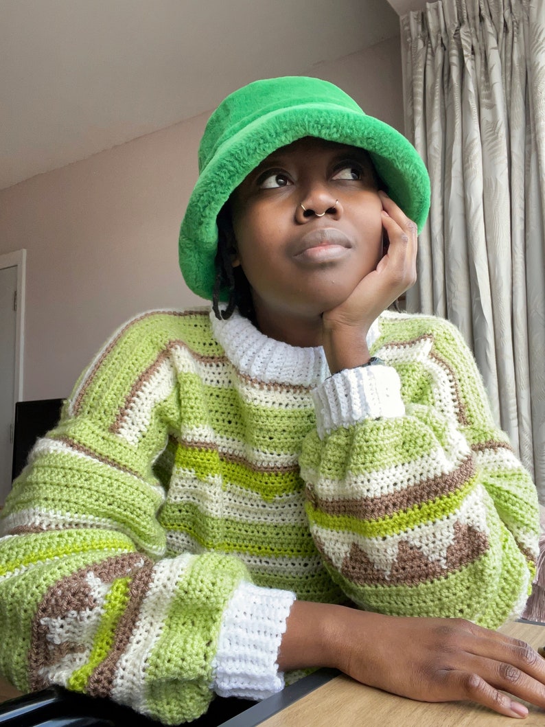 May include: A person wearing a green fuzzy bucket hat and a green, white, and brown striped crocheted sweater.
