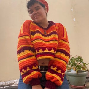 May include: A cropped, multi-colored crochet sweater with a striped pattern in shades of orange, yellow, and maroon. The sweater has a loose fit and a slightly flared sleeve.