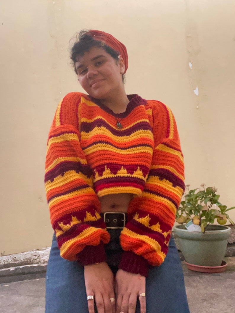 May include: A cropped, multi-colored crochet sweater with a striped pattern in shades of orange, yellow, and maroon. The sweater has a loose fit and a slightly flared sleeve.