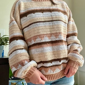 May include: A crocheted sweater with brown, beige, and white stripes and a geometric pattern. The sweater has a relaxed fit and long sleeves.