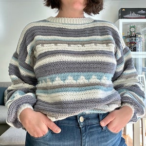 May include: A striped crocheted sweater with a white, gray, blue, and brown color scheme. The sweater has a round neckline and long sleeves.