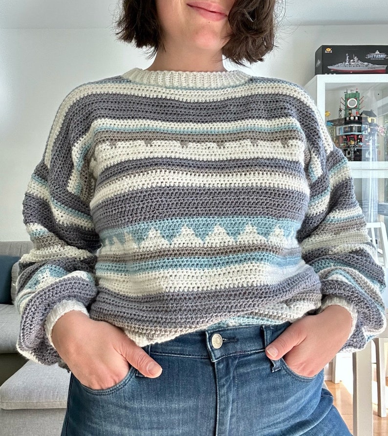May include: A striped crocheted sweater with a white, gray, blue, and brown color scheme. The sweater has a round neckline and long sleeves.