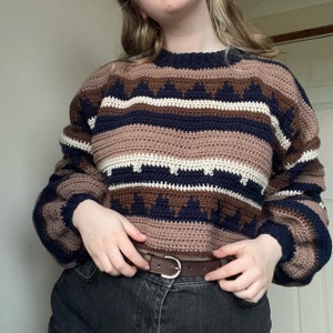 May include: A brown, white, and navy blue striped crocheted sweater with a geometric pattern. The sweater has a round neckline and long sleeves.