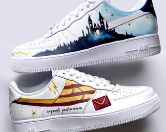 May include: A pair of white sneakers featuring custom hand-painted designs. One shoe displays a dark blue castle silhouette against a light blue sky with gold stars. The other shoe features a red and gold striped scarf, a red envelope, and the text expecto patronum.