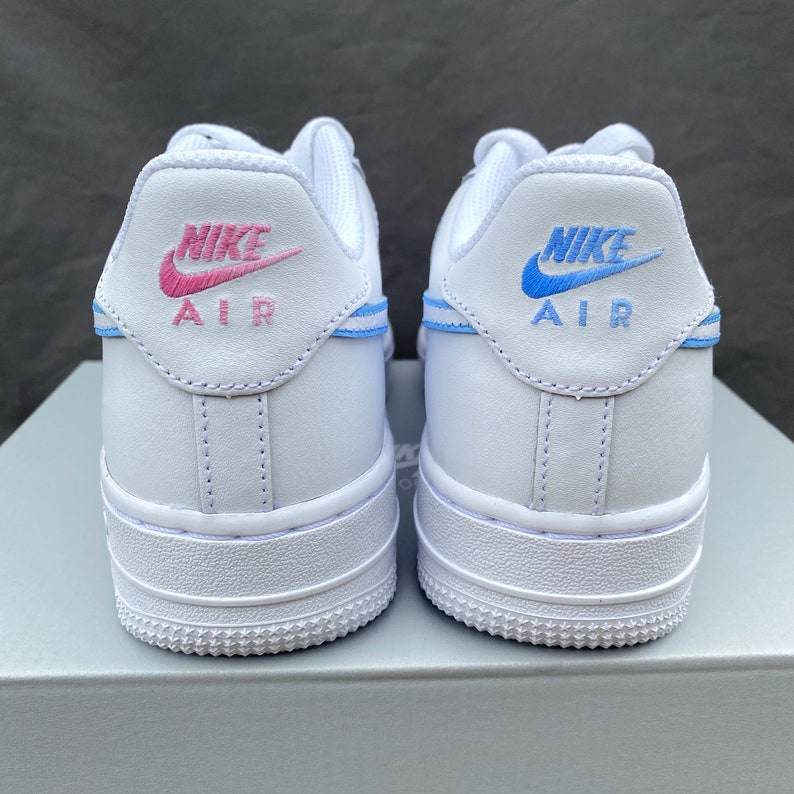 K&ouml;nnte beinhalten: Ein Paar wei&szlig;e Nike Air Force 1 Sneaker mit blauen und rosa Akzenten. Der linke Schuh hat einen rosa Nike Swoosh und den Text "AIR", w&auml;hrend der rechte Schuh einen blauen Nike Swoosh und den Text "AIR" hat.