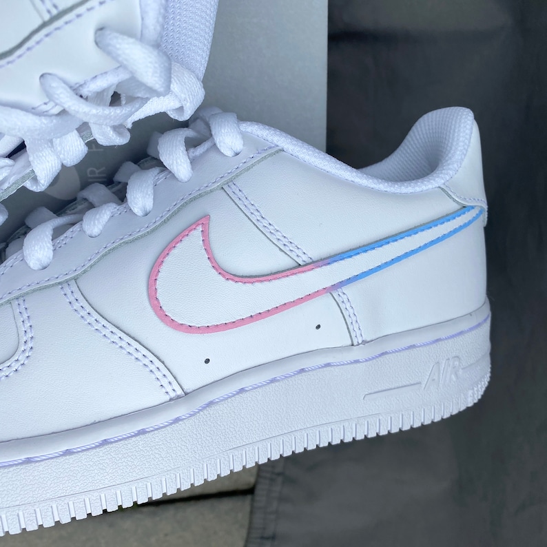 K&ouml;nnte beinhalten: Wei&szlig;e Leder-Sneaker mit niedrigem Schaft und einem pinkfarbenen und blauen Swoosh-Logo. Die Sneaker haben eine wei&szlig;e Gummisohle mit einem hellblauen "AIR"-Logo.