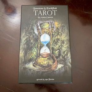 May include: A black box with a dark green and brown illustration of an hourglass with skeletal hands holding it. The text on the box reads "Bonestone & Earthflesh TAROT By Avalon Cameron Artwork by Ana Tourian".