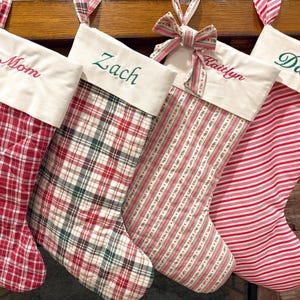 Puede incluir: Cuatro calcetines navideños con puños blancos que llevan bordados los nombres Mom, Zach, Kaitlyn y Dad. Los calcetines presentan varios patrones en rojo y blanco, incluyendo cuadros acolchados, cuadros verdes y rojos, rayas florales y rayas finas.