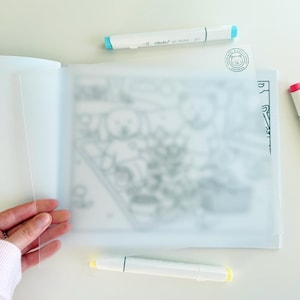 May include: An open art book with a clear plastic overlay, featuring a partially visible black and white illustration. Three white dual-tip markers with colored ends and caps in blue, pink, and yellow are scattered around the book.