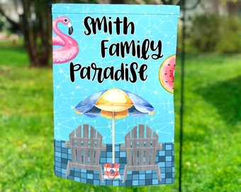 May include: A blue and white garden flag with a pool scene. It features a pink flamingo float, two lounge chairs, an umbrella, and a watermelon. The text "Smith Family Paradise" is written in black.