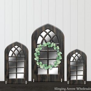 May include: Three wooden arched window frames with a faux wood finish. The largest frame has a eucalyptus wreath hanging on it. The frames are all different sizes and are standing on a dark surface.  Slinging Arrow Wholesale is printed at the bottom of the image.