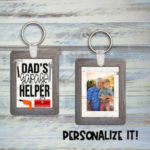 Puede incluir: Dos marcos de fotos de madera con llaveros. El marco de la izquierda tiene un fondo blanco con texto negro que dice "Dad's garage helper" y una caja de herramientas roja, un taladro y un martillo. El marco de la derecha tiene una foto de dos personas sonriendo.