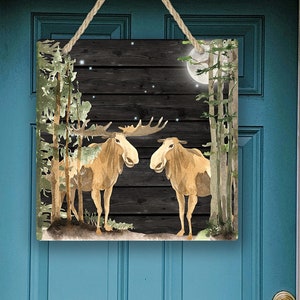 Op de afbeelding: Een houten bord met een aquarel-illustratie van twee elanden die 's nachts in een bos staan. De achtergrond is een donkere houten plank met volle maan en sterren.