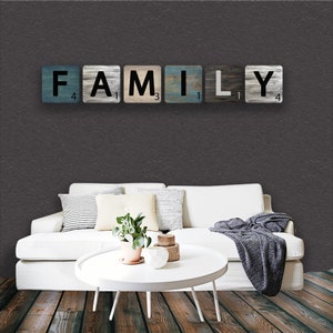 May include: A white couch with pillows and a throw blanket sits in front of a dark gray wall. The word "FAMILY" is spelled out in black letters on wooden tiles with a blue and gray paint design. The tiles are arranged on the wall above the couch.