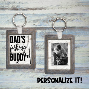 Puede incluir: Dos marcos de fotos de madera con llaveros. El marco de la izquierda tiene un gráfico en blanco y negro de una caña de pescar y el texto "DAD'S fishing BUDDY". El marco de la derecha tiene una foto en blanco y negro de un padre y un hijo pescando.