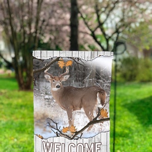 Pode incluir: Um sinal de madeira branco com um visual desgastado que apresenta um veado de cauda branca em um ambiente de floresta nevada. O sinal diz "Welcome The Millers" em texto preto.