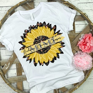 Peut inclure: T-shirt blanc avec un motif de tournesol jaune. Le tournesol a un motif léopard noir et brun. Un ruban doré avec le texte "Nicholson Kima Cooper" est enroulé autour du tournesol.