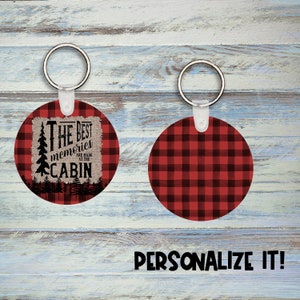 May include: Two round keychains with a red and black buffalo plaid pattern. One keychain has a burlap tag with the text "The Best Memories Are Made At The Cabin" and a small pine tree graphic.