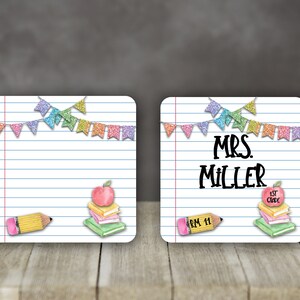 以下が含まれることがあります： 白い背景と青い線でできた正方形の先生の名前タグが2つ。それぞれのタグの上には、カラフルな旗の列があります。左側のタグには、黄色の鉛筆と赤いリンゴが乗った本の束があります。右側のタグには、「MRS. MILLER」という文字と、赤いリンゴが乗った本の束があり、「1ST GRADE」と書かれています。