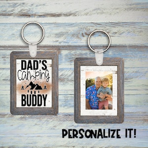 Puede incluir: Dos llaveros rectangulares con un marco de aspecto de madera envejecida. Un llavero tiene el texto "DAD'S CAMPING BUDDY" con gráficos de montañas y fogatas. El otro llavero muestra una foto de un hombre y un niño. El texto "¡PERSONALÍZALO!" está debajo.