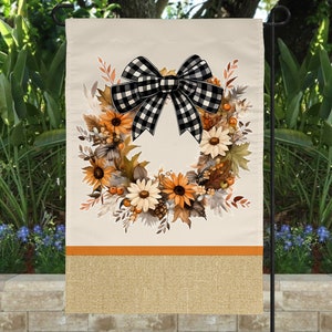 May include: A fall-themed garden flag with a wreath of sunflowers, leaves, and berries. A black and white checkered bow is tied at the top of the wreath. The flag has a burlap border with an orange stripe.