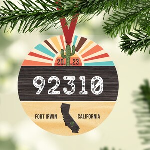 Puede incluir: Un adorno navideño redondo de madera con un diseño retro de rayos de sol, un cactus y el texto "92310 Fort Irwin, California".
