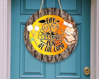 Könnte beinhalten: Ein rundes Holzschild mit gelbem Hintergrund und brauner Baumrindenstruktur. Das Schild sagt "Life is s'more fun at the camp" mit einem Cartoon-Bild von zwei lächelnden Marshmallows und einem Marshmallow auf einem Stock.