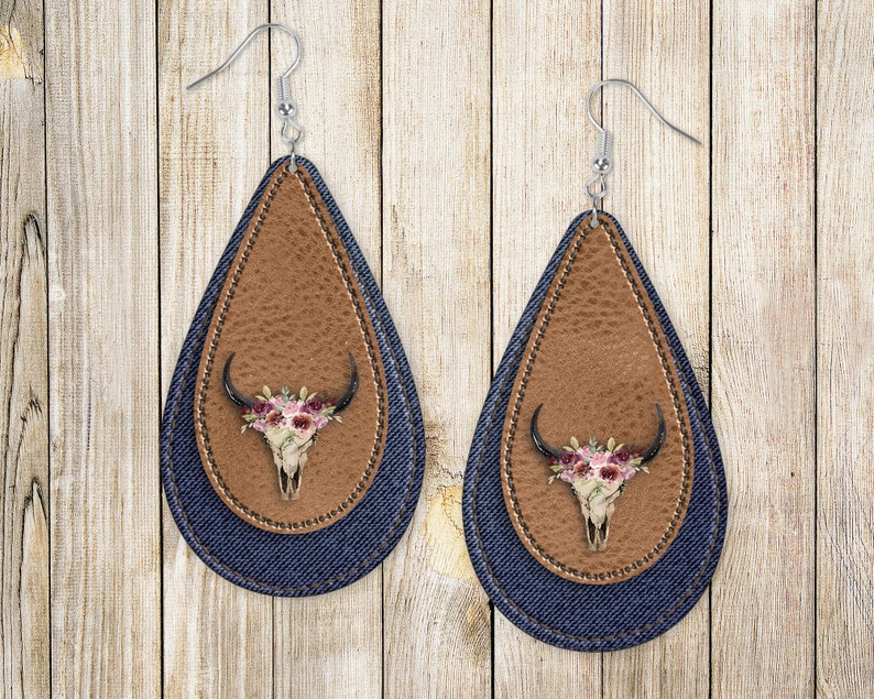 May include: A pair of teardrop-shaped earrings with a brown leather and denim design. The earrings feature a floral skull design with pink and white flowers.