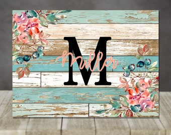 May include: A wooden sign with a distressed turquoise and brown background. A large black letter "M" is centered, with the name "Miller" in pink script below. Floral watercolor accents in pink, blue, and green adorn the corners.